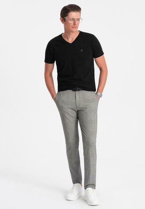 Uomo che indossa una maglietta nera a V, pantaloni grigi a quadri, sneakers bianche, occhiali e un orologio da polso, in piedi con le mani nelle tasche.