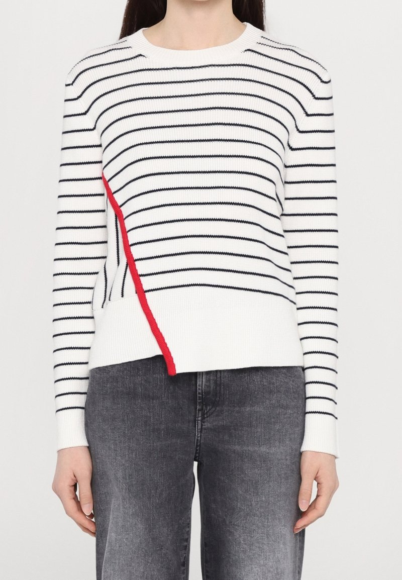 Pull en maille blanche à rayures horizontales noires, avec un accent rouge sur le côté. Le modèle comprend un col rond et des poignets côtelés.