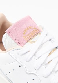 Bílé kožené tenisky s bílými tkaničkami a růžovou semišovou jazykovou štítkem s embosovaným zlatým textem v němčině.