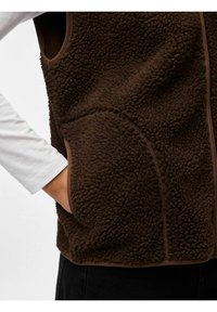 Rudos vilnos liemenė su tekstūruotu paviršiumi, užtrauktuku priekyje ir šoninėmis kišenėmis. Apdailos kraštai kontrastuoja su šviesesne ruda spalva. Po ja vilkima ilgomis rankovėmis marškiniai.