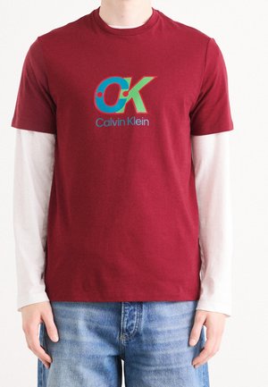 Personne portant des vêtements superposés avec un t-shirt rouge Calvin Klein par-dessus une chemise blanche à manches longues et un jean bleu.