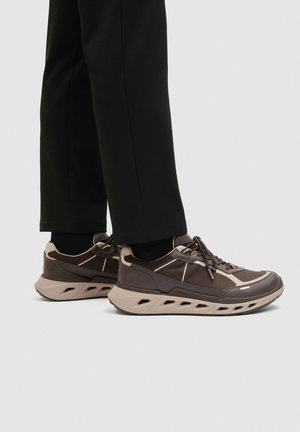 Braune und beige Sneaker mit perforierten Sohlen, getragen zu schwarzen Hosen und schwarzen Socken, seitlich auf hellem Hintergrund gezeigt.