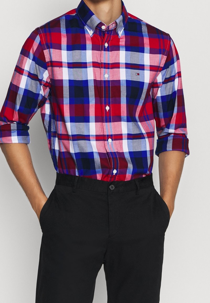 Camisa de cuadros roja, azul y blanca con botones, cuello, mangas largas remangadas y botones blancos; combinada con pantalones negros.