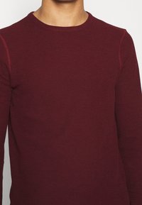 Bordowy dzianinowy longsleeve z okrągłym dekoltem i kontrastującymi szwami na ramionach. Miękka faktura i dopasowany krój.