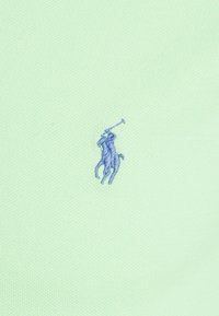 Modrý vyšívaný loga polo hráče na koni na světlém zeleném texturovaném pozadí.