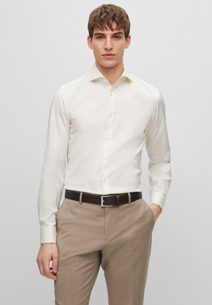 Chemise en beige clair avec un col pointu, des manches longues et une fermeture à boutons, accompagnée d'un pantalon beige et d'une ceinture marron foncé.