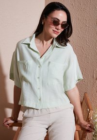Blusa verde chiaro a maniche corte con vestibilità rilassata, chiusura frontale con bottoni e due tasche sul petto. Tessuto morbido e leggero con una sottile texture.
