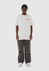 Hvit oversized t-skjorte med rød grafisk tekst, kombinert med mørkegrå slitte jeans med store hull og hvite sneakers.
