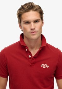 Polo rouge en coton, doté d'un col à deux boutons, avec le logo brodé « SUPER DRY » en blanc, et des manches courtes à la texture lisse.
