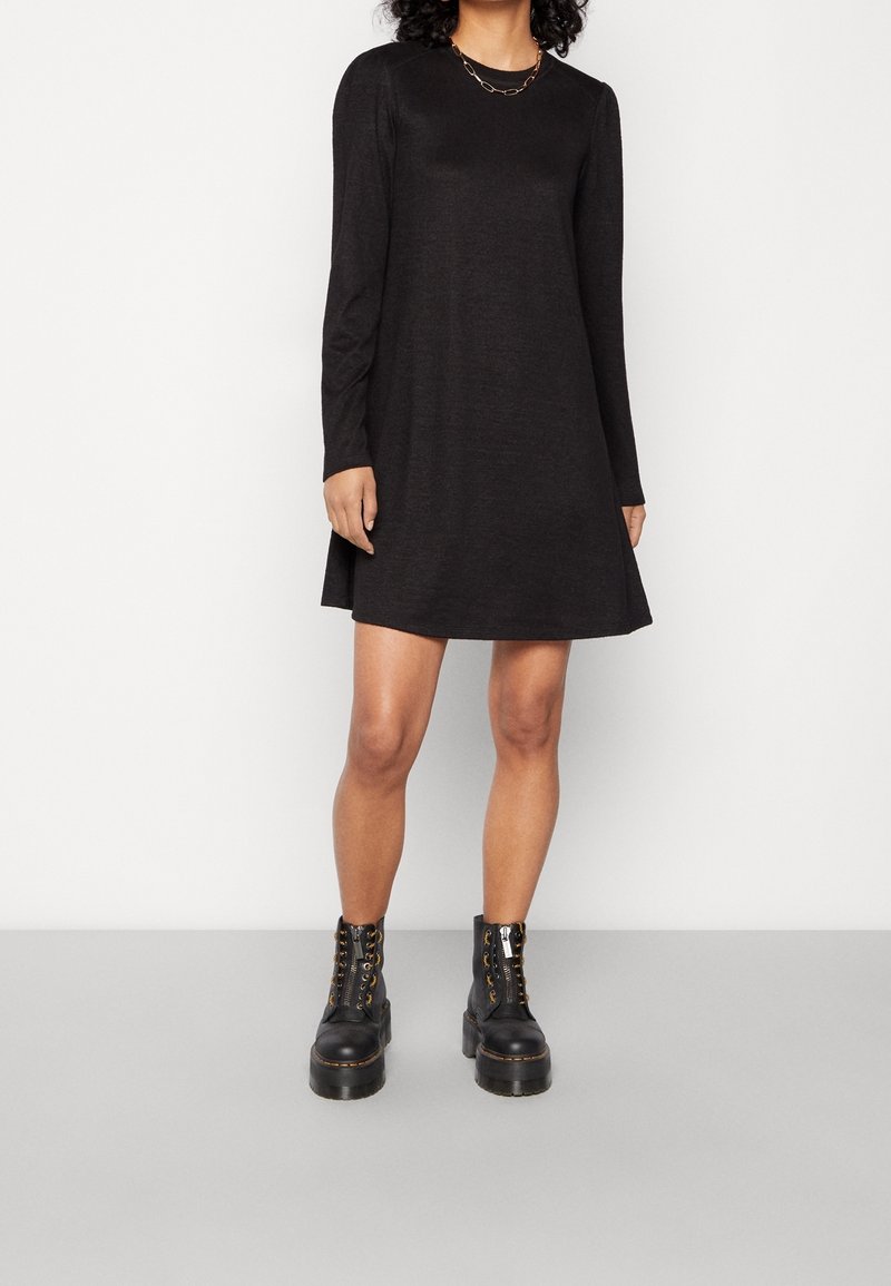 Femme portant une robe noire à manches longues et des bottines à plateforme noires lacées, debout devant un fond blanc uni.