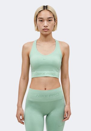 Jeune personne aux cheveux blonds courts, portant un soutien-gorge de sport Nike Pro vert clair et un legging taille haute assorti, debout, face à l'avant.