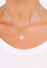 Colliers argentés superposés avec une chaîne fine et un pendentif circulaire texturé. Les chaînes sont délicates, mettant en valeur un chemisier blanc.