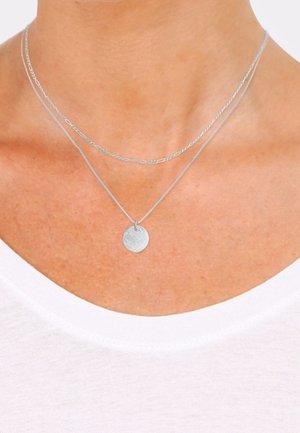 Colliers argentés superposés avec une chaîne fine et un pendentif circulaire texturé. Les chaînes sont délicates, mettant en valeur un chemisier blanc.