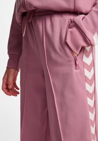 Roze sportieve broek gemaakt van een gestructureerde stof. Kenmerken zijn onder andere zijzakken met ritsen en een wit chevronpatroon langs het been.