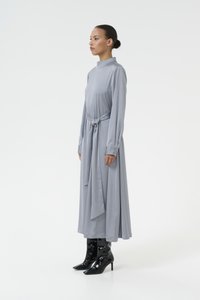 Robe grise longue avec un col montant, cintrée à la taille. Elle présente de longues manches et une texture lisse, assortie à des bottes noires à talons hauts et brillantes.