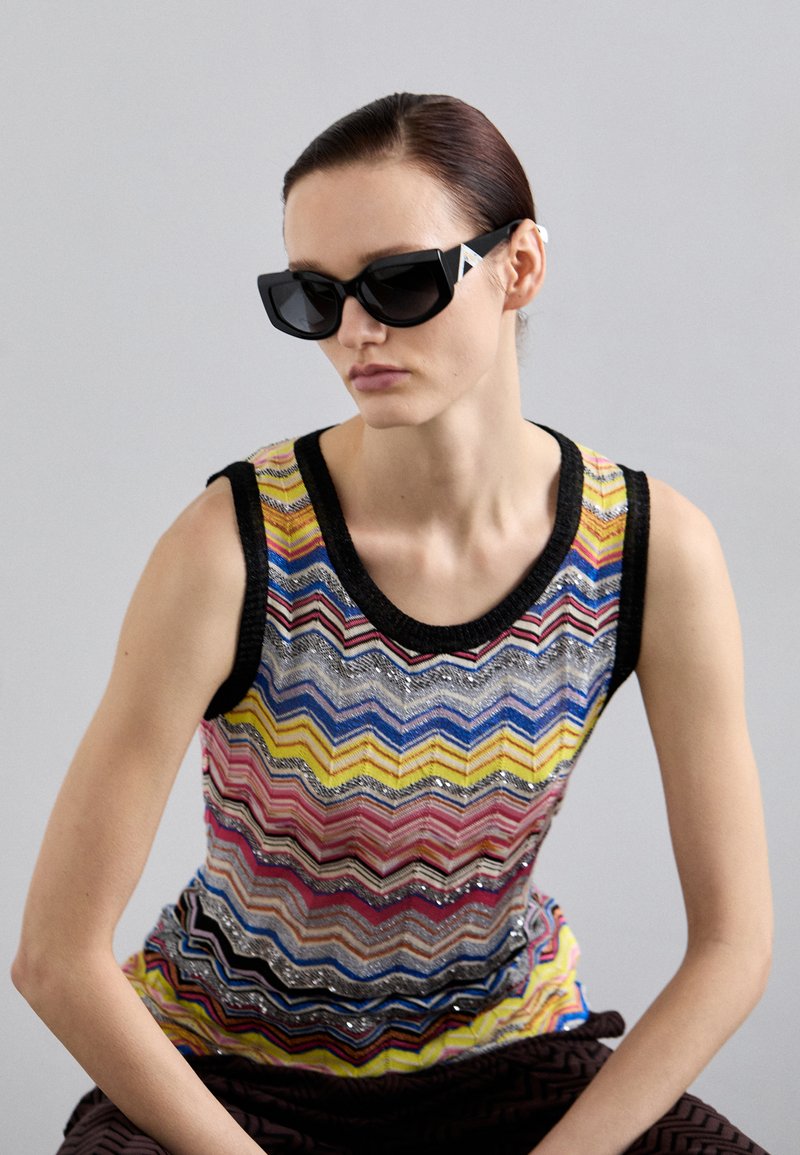 Multicolored sleeveless top with a zigzag pattern, embellished with sequins, featuring a black neckline. Worn with black angular sunglasses.