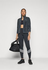 Zwarte sportjack met een textuurpatroon, gecombineerd met zwarte legging met grijze accenten. Model houdt een zwarte sporttas vast.