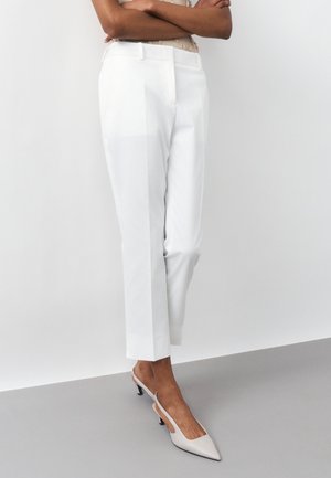 Άτομο που φοράει λευκό, ραμμένο στα μέτρα, παντελόνι μήκους έως τον αστράγαλο και λευκά slingback ψηλοτάκουνα παπούτσια με μυτερή μύτη, στέκεται με σταυρωμένα τα χέρια μπροστά σε απλό φόντο.