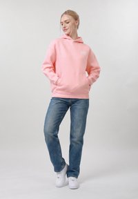 Femme debout, les mains dans les poches, portant un sweat à capuche rose, un jean bleu et des baskets blanches, sur un fond clair uni.