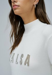 Vrouw in een witte geribde coltrui met rhinestone letters die "MALISA" spellen, met zilveren oorbellen in de vorm van ringen, weergegeven van nek tot borst.