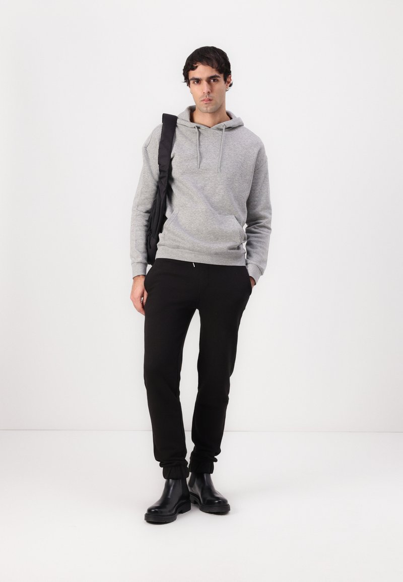 Sweatshirt gris à capuche avec une poche kangourou, associé à un pantalon de jogging noir et des bottines noires brillantes, le tout porté avec un sac noir à l'épaule.