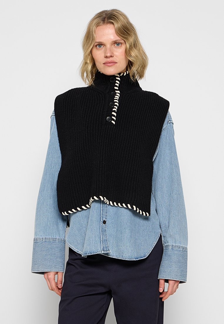 Schwarze Strickweste mit hohem Kragen, ausgestattet mit drei Knöpfen und weißem Saum, getragen über einem hellen Jeanshemd.
