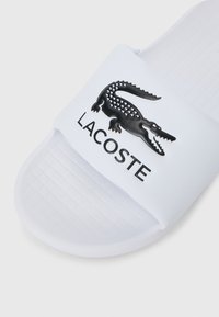 Vita sandaler i vitt med en texturerad rem som har en svart krokodil-logotyp och texten "LACOSTE" i svart under, platt sula med rutmönster.