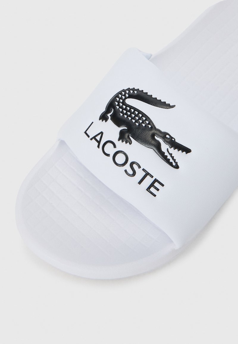 Weiße Rutschsandale mit einem strukturierten Riemen, einem schwarzen Krokodil-Logo und dem Schriftzug "LACOSTE" in Schwarz darunter, flache Sohle mit Rillenmuster.