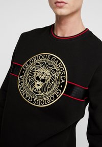 Sudadera negra con un gran emblema de león dorado, letras "GLORIOUS GANGSTA" y franjas de acento rojas y negras en las mangas y el cuello.