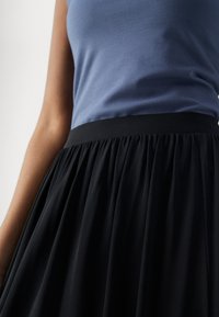 Detailní záběr osoby v modrém topu bez rukávů, který je zasunutý do černé plisované sukně s vysokým pasem.