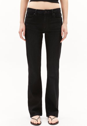 Personne portant un jean noir taille haute à jambes droites et des sandales noires en tongs, debout devant un fond blanc.