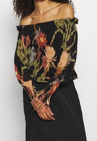 Donna con una blusa nera off-shoulder decorata con una stampa floreale colorata e maniche lunghe a sbuffo, abbinata a una gonna nera plissettata.