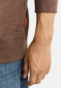 Maglione marrone a coste con scollo rotondo, caratterizzato da una piccola etichetta in pelle sul lato. Manica aderente e jeans casual blu denim visibili.