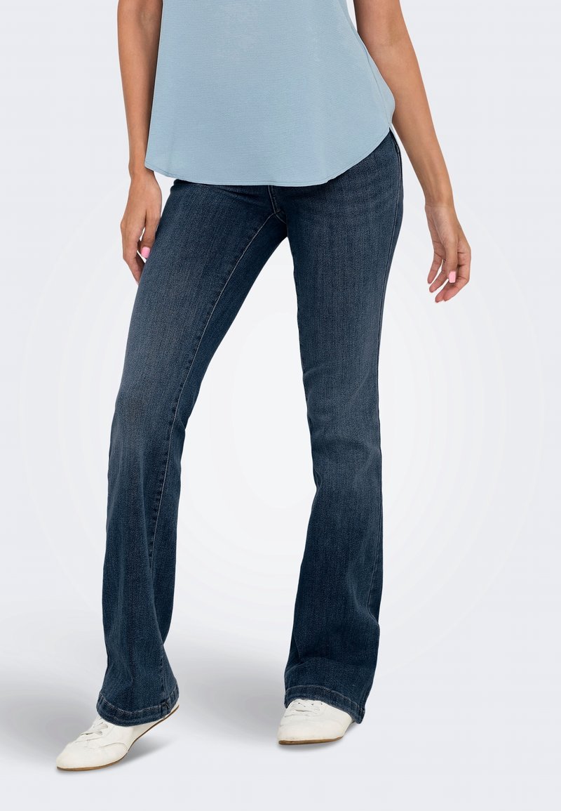 Femme portant un jean bootcut bleu foncé, un haut sans manches bleu clair et des baskets blanches, debout dans une posture détendue.