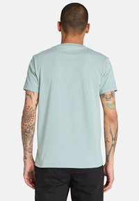 Ljusblå kortärmad t-shirt med rund hals och en slät textur, med synliga tatueringar på armarna hos personen som bär den.