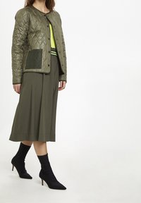 Chaqueta verde acolchada con botones a presión, combinada con una falda oliva fluida y botines negros de punta. Acento en una blusa de color verde brillante.