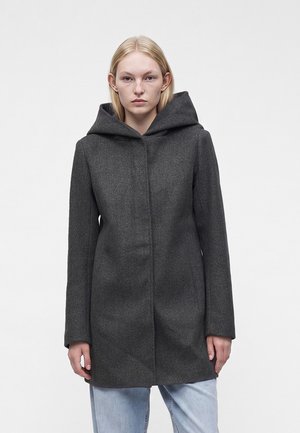 Femme portant un manteau à capuche gris foncé, debout devant un fond clair uni.
