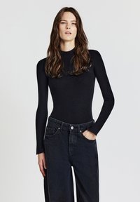 Schwarzes, geripptes Langarmoberteil mit hohem Kragen, kombiniert mit weit geschnittenen, dunklen Jeans aus Denim mit frontalen Taschen und einem Knopfverschluss.