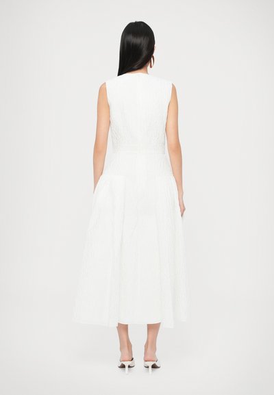 Bela obleka brez rokavov s teksturiranim materialom, oprijetim zgornjim delom in krilom v obliki črke A. Poudarjen pas, stranski žepi in minimalno vidni šivi.