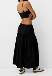 Schwarzer, gestufter Maxirock mit elastischem Bund, kombiniert mit einem schwarzen Cropped-Top. Das Model trägt weiße Sneakers mit schwarzen Akzenten.