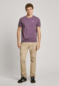 Camiseta de algodón morada con escote redondo y logo blanco, combinada con chinos beige y zapatillas negras con acentos blancos.