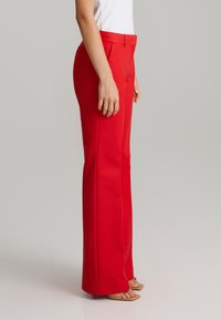 Pantalones rojos con un corte ajustado, textura lisa, cinturilla de altura media y bolsillos laterales, combinados con una blusa blanca sencilla y sandalias beiges.