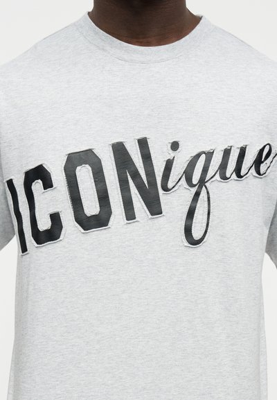 Άτομο που φοράει ανοιχτό γκρι μπλουζάκι με τη λέξη "ICONique" σε μεγάλα μαύρα γράμματα στο στήθος.