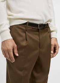 Bruine broek met voorplooien, gecombineerd met een crèmekleurige trui. Een zwarte leren riem met een zilveren gesp accentueert de outfit.