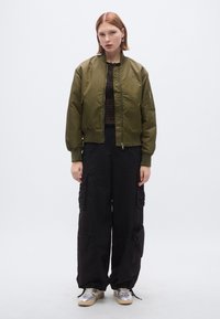 Πράσινο μπουφάν μπούμερ, με φερμουάρ, με ριμπ μανσέτες. Μαύρο παντελόνι cargo με πολλαπλές τσέπες. Ασημένια αθλητικά παπούτσια με υφή.