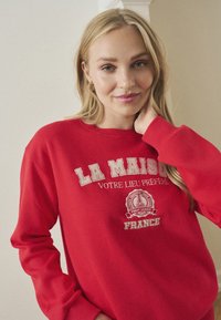 Felpa rossa con girocollo, maniche lunghe e testo ricamato in beige: "LA MAISON, VOTRE LIEU PRÉFÉRÉ, FRANCE." Tessuto morbido.