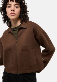 Veste brune zippée en tissu texturé, coupe oversize, et deux poches avant. Elle possède un col et est portée sur une chemise verte.