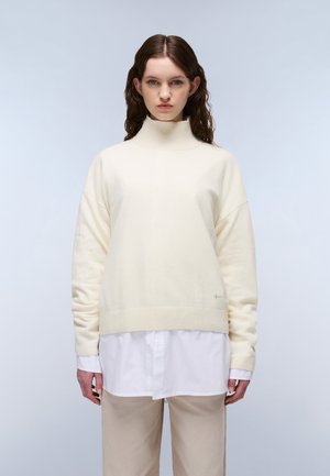 Ung kvinde med brunt bølget hår iført cremefarvet rullekravetrøje over hvid skjorte med knapper og beige bukser, stående mod en ensfarvet baggrund.
