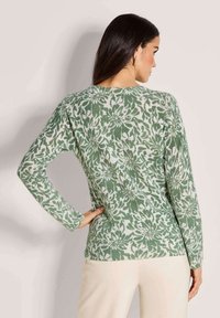 Frau mit langen dunklen Haaren, die einen grün-weißen Pullover mit Blumenmuster trägt, mit einer Hand auf der Hüfte stehend und von hinten blickend.