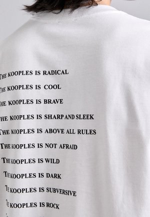 Osoba ubrana w białą koszulkę z czarnym tekstem wymieniającym frazy zaczynające się na "The Kooples is,", w tym radykalny, fajny, odważny, bystry, dziki i podważający.